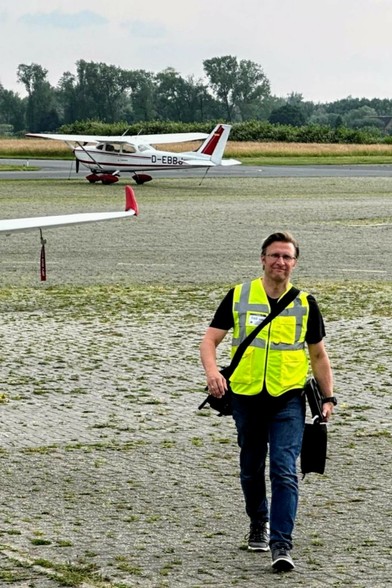 man with yellow west walking on the apron of an airfield, headset case in the left hand.