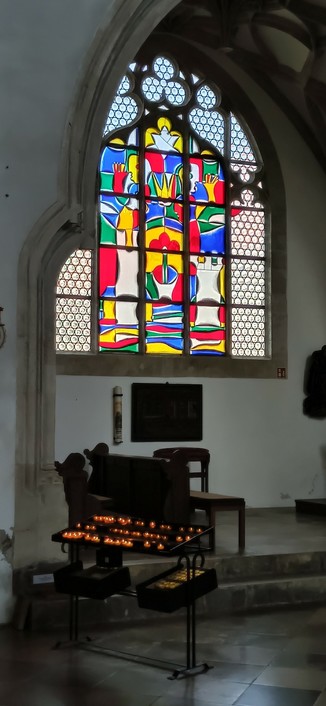Farbiges Kirchenfenster in gotischem Rahmen  und ein Kerzen Regal im Vordergrund, auf dem viele kleine Kerzen brennen