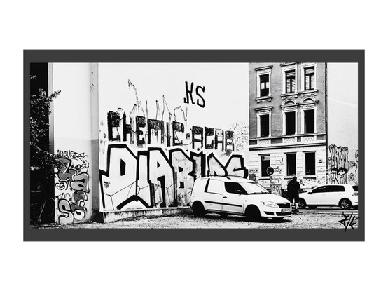 Schwarz-Weiß Foto von einem Graffitischriftzug der Ultragruppe Diablos von Chemie Leipzig an einer hellen Hauswand auf einem Parkplatz. Davor steht ein weißer Kaddy Transporter.
