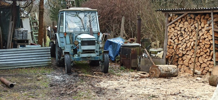 Alter Traktor in einem Bayernhof abgestellt, rechts daneben ein überdachter Holzstapel mit Sägespänen vor dem Stapel vom HOlzmachen, linke vom Traktor ein Wellblechstreifen und allerlei alte Gerätschaften.