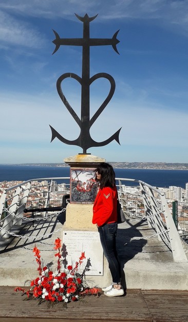 Eine Frau steht vor einem Denkmal der ertrunkenen Fischer von Marseille. In der Ferne das Meer, dahinter die Küste und einige Häuser.