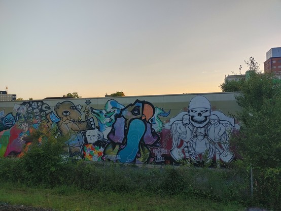 buntes Graffiti mit Monsterfiguren an einer Bahnhofswand hinter dem Grünstreifen am Gleis