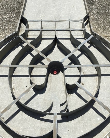 Mein symmetrisch aufgebautes Foto zeigt das Ziellabyrinth einer Minigolfbahn. Man erkennt verschiedene durch Metallleisten definierte Wege, die der Ball nehmen kann, um im Loch zu landen. Die innerste Struktur erinnert an ein Herz, der Ball im Loch ist rot.