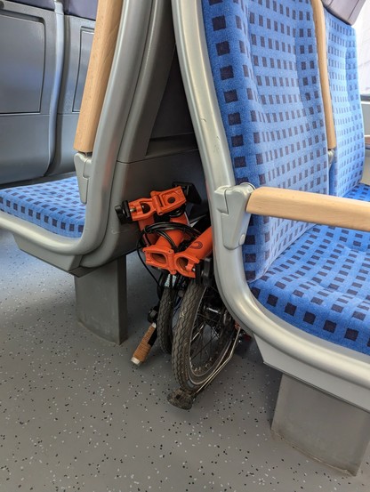 ein gefaltetes Rad ist Marke Brom in der Farbe fire coral welches zwischen zwei Sitzen, die Lehne an Lehne angeordnet sind, in einem Regionalzug steht.