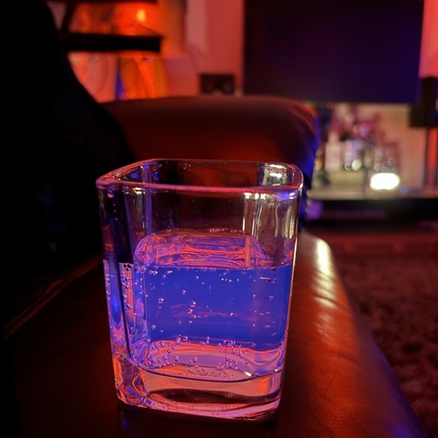 a glass on a chair, the room is bathed in red light apart from some out of focus candles in the background; the drink in the glass is glowing blue