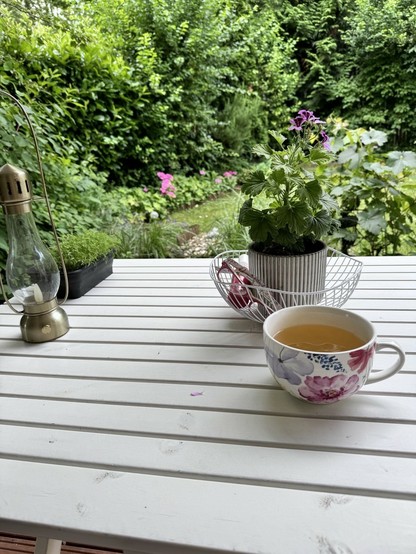 Weißer Holztisch , darauf eine große geblümte Tasse mit Grüntee, dahinter einKorb mit ner Pflanze drin. Im Hintergrund Viel Grun Kit ein paar Hortensienblüten. 