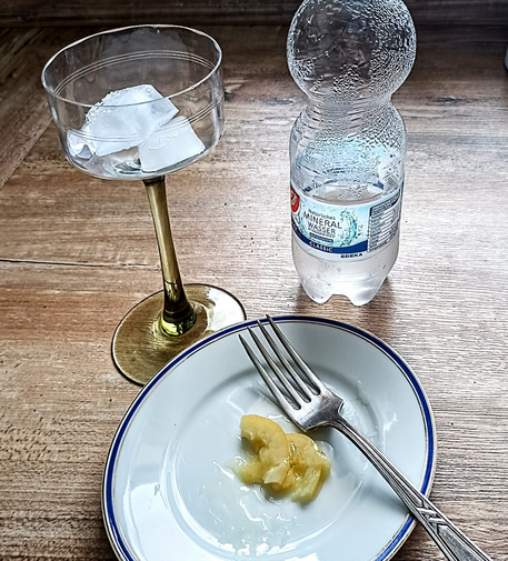 auf einem Teller einige Stücke Salzzitrone und eine Gabel, dahinter ein Weinglas mit Eiswürfeln und eine Flasche Mineralwasser