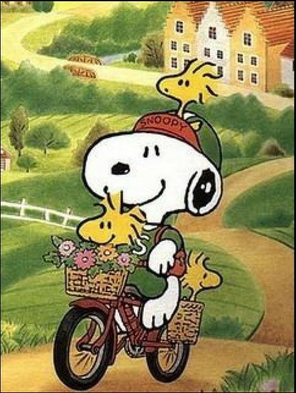 Snoopy fährt auf einem Fahrrad durch eine liebliche Landschaft. Auf seiner Basecap sitzt Woodstock, in den Fahrrad Körben vorne und hinten zwei von Woodstocks Freunden