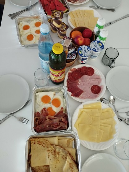 Ein Frühstückstisch gedeckt mit Pfannkuchen, Käse, Spiegeleier, Speck, Wurst, Saft, Melone, Nektarine, verschiedenen Joghurt