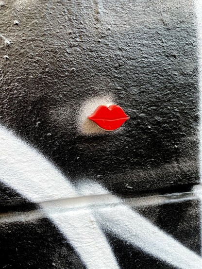 Nahaufnahme einer strukturierten schwarzen Wand mit weißen Graffiti-Linien. In der Mitte sind zu einem Kuss zugespitzte rote Lippen zu sehen. 
Streetart im der Schanze in Hamburg. 