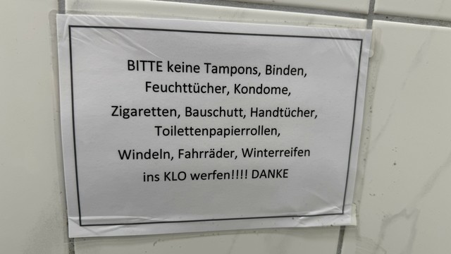 A sign in German on a tiled wall listing items not to be flushed down the toilet, including tampons, sanitary pads, wet wipes, condoms, cigarettes, construction debris, towels, toilet paper rolls, diapers, bicycles, and winter tires.