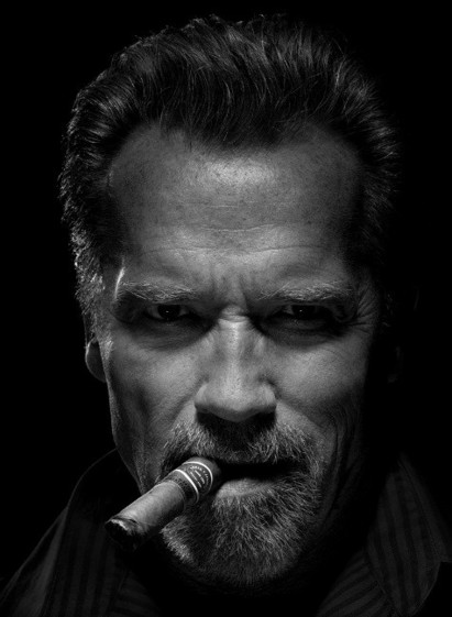 Schwarz-weiß Portrait von Arnold Schwarzenegger mit Zigarre im rechten Mundwinkel.