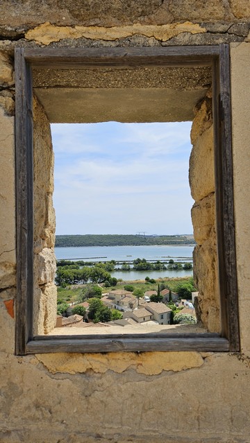 Unverglastes Fenster mit einfachem schmalem Eichenholzrahmen in morbide Steinmauer eingefasst, mit Ausblick auf mediterrane Häuser, einen See und Wald 