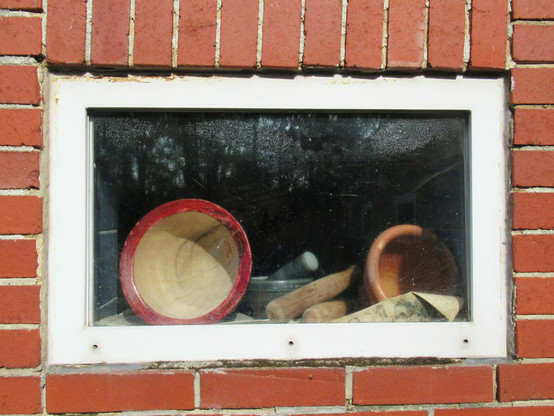 Ein kleines, rechteckiges Fenster in einer Fassade aus rotem Klinker. Durch die Scheibe sind ein leerer Topf und eine leere Schale sowie Griffe von Werkzeug erkennbar.
