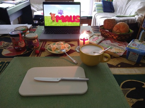 Ein gedeckter Frühstückstisch mit Banane, Brötchen, Käse, Marmelade, Honig, Cappuccino. Eine Kerze brennt. Im Hintergrund läuft die Sendung mit der Maus.