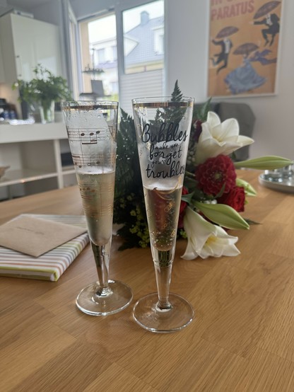 Two champagne flutes with bubbly drinks, one with musical notes and the other with a quote. A wrapped gift and a bouquet of flowers are on the table. Blurred background includes kitchen and a poster on the wall.