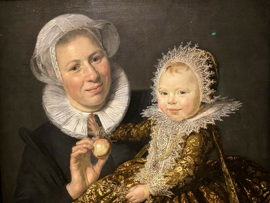 Das Gemälde zeigt ein kostbar gekleidetes Kind auf dem Arm einer lächelnden Amme. 