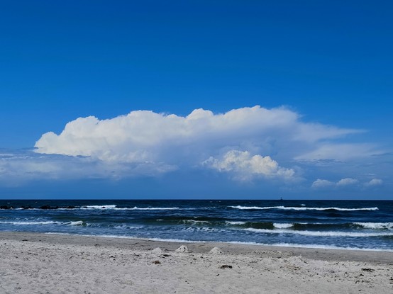 Stahlblaues Meer mit weißen Wolken am Horizont 
Man erkennt Wellen und Gischt
Der Strand ist leer