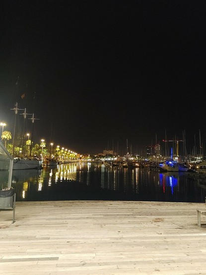 Lichter der Boote und Häuser am Hafen spiegeln sich im schwarzen Wasser unter schwarzem Himmel, im Vordergrund der von Straßenlaternen beleuchtete Holzboden der Rambla del Mar