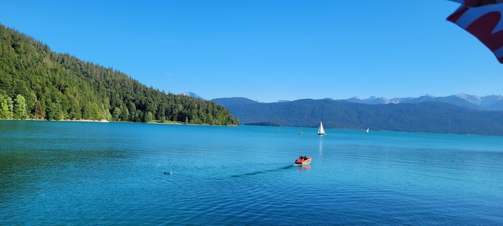 Der Walchensee mit einem Tretboot und weiter hinten ein Segelboot. Im Hintergrund Berge, von links ein bewaldeter Hügel