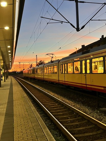 Beleuchtetes Bahngleis am frühen Morgen, am gegenüberliegenden Gleis steht eine Straßenbahn, der Himmel leuchtet im Morgenrot