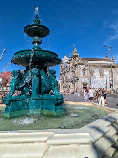 im Vordergrund ein Brunnen mit 4 geflügelten Löwen vor 2 nebeneinander stehenden Kirchen in Porto im Hintergrund 