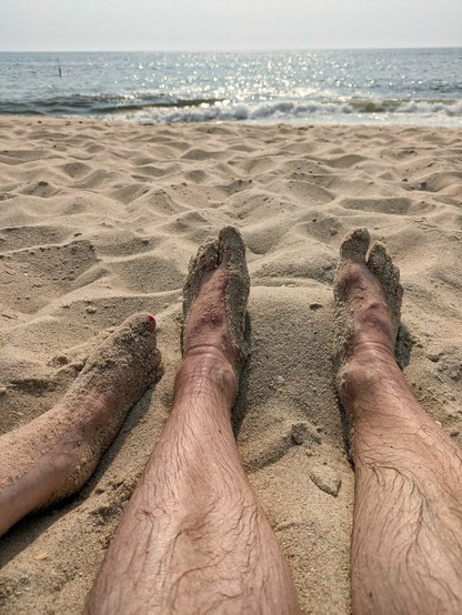 drei nackte Beine liegen im Sand und dahinter unscharf Wellen.sonnig