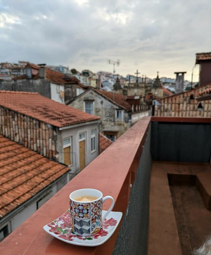 eine Espressotasse steht auf der Blecheinfassung von der Balkonmauer. Dahinter unscharf eine große Stadt mit vielen verschiedenen alten kleinen Dächern und ein paar Kirchtürmen 