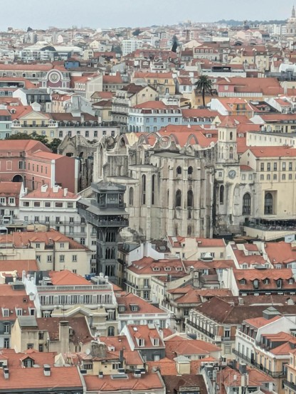 Blick auf die Altstadt von Lissabon mit vielen kleinen Häusern mit roten Ziegeldächern. Ein dunkler großer Aufzug und die Ruine einer großen Kirche 