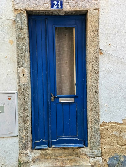 eine blaue Haustür, 1/4 zu 3/4 geteilt und im breiten Teil ein längliches Fenster mit beigem Vorhang