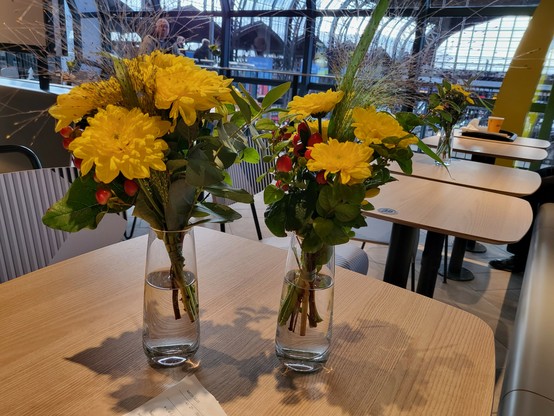 Auf einem hellbraun Tisch stehen zwei Glasvaden mit gelben Chrysanthemen und roten Ilex. Im Hintergrund die Halle des Hamburger Hauptbahnhofes