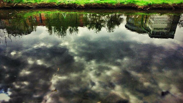 Spiegelung im Wasser zeigt einen Himmel mit dicken schwarzen Wolken, aber auch ein Haus und mehrere Bäume