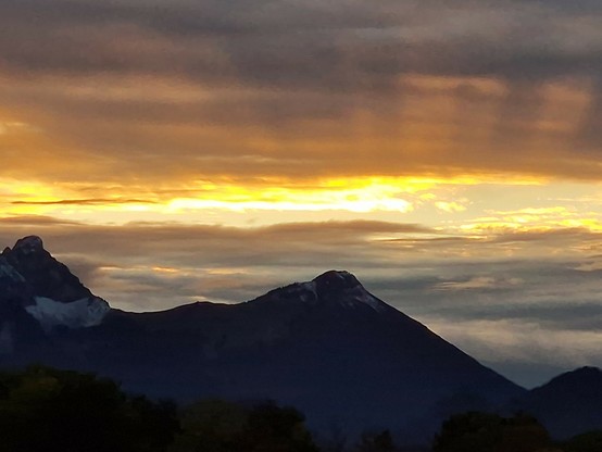 Sonnenuntergang hinter Wolken, gelb-orangefarbene Streifen und dunkle Strahlen hinter dunklen Bergen.