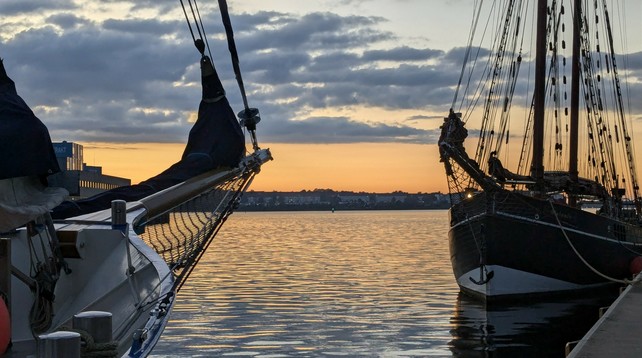 Sunset. Grey blues and life Mon oranges. A harbour. The bowsprits and forward rigging of two small schooners in the failing light.