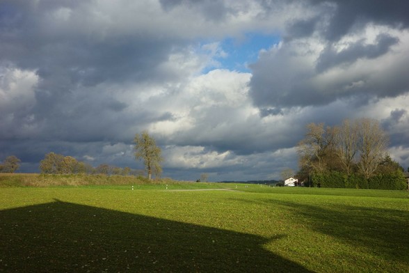 dunkle und helle Wolken haben ein kleines Loch, durch das blauer Himmel zu sehen ist.nDie Sonne erzeugt auf der grünen Wiese einen Schatten vom Haus 