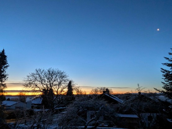 blauer Himmel mit einem kleinen Mond über bunten Streifen am Horizont.nSchnee auf den Dächern der Häuser und im Garten 