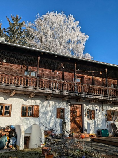 hinter einem alten Bauernhaus mit Holzaufbau ist eine komplett weiß angereifte Birke und blauer Himmel zu sehen 