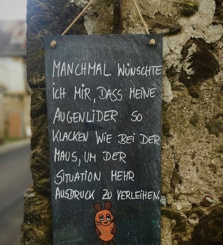 Bild zeigt ein schwarz-weißes Schild, das an einer steinernen Wand befestigt ist. Das Schild ist aus Naturstein und hat eine matte Oberfläche. Es ist mit weißer Kreide geschrieben, die auf dem dunklen Hintergrund gut sichtbar ist. Der Text auf dem Schild lautet: 