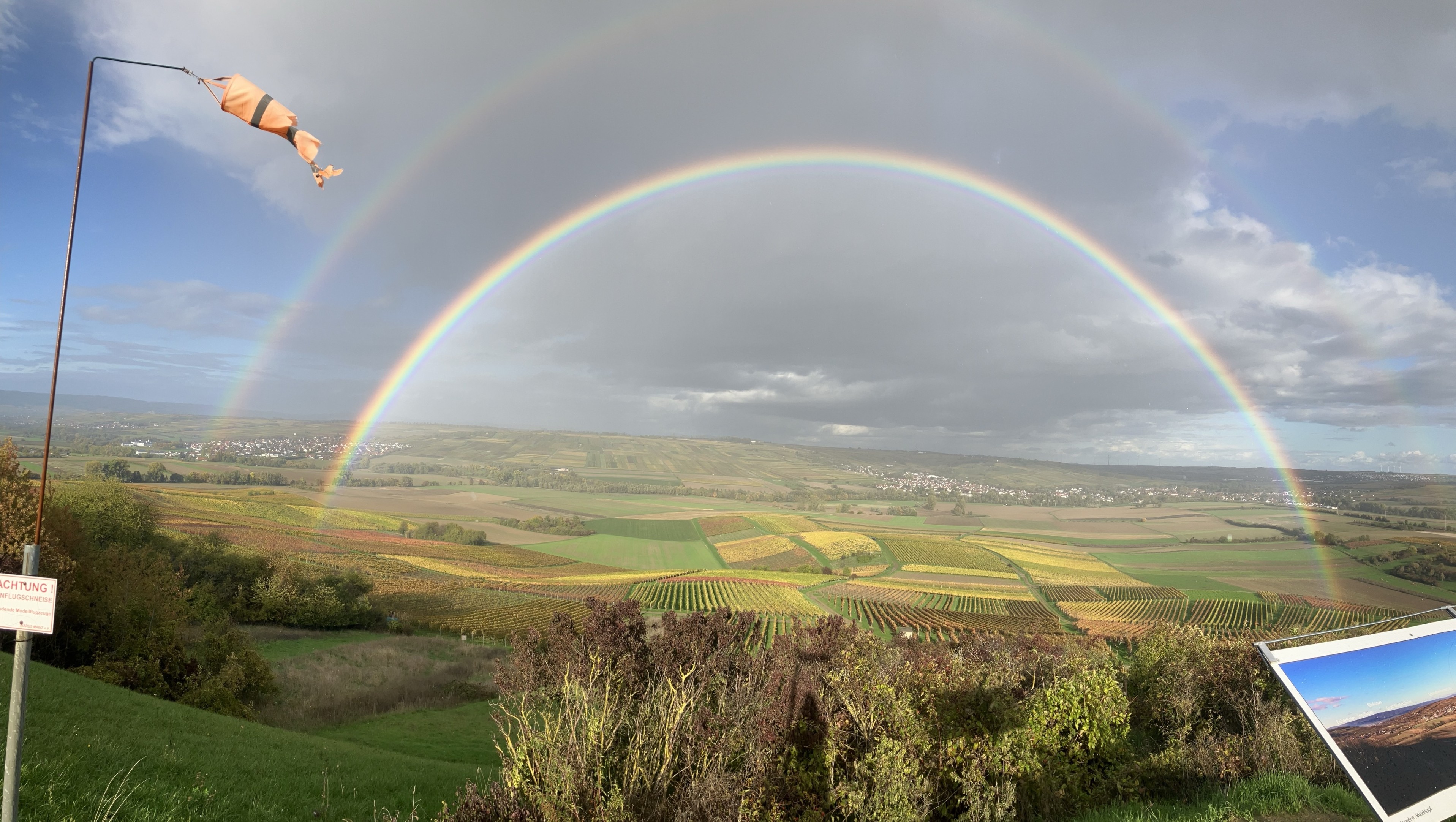 Ein Panoramablick auf sanfte grüne und gelbe Felder unter einem dramatischen Himmel, mit einem doppelten Regenbogen, der sich über die Landschaft wölbt. Es gibt eine Flagge auf einer Stange und einen Wegweiser im Vordergrund, was der Einstellung Kontext hinzufügt. Ein Panoramablick auf sanfte grüne und gelbe Felder unter einem dramatischen Himmel, mit einem doppelten Regenbogen, der sich über die Landschaft wölbt. Es gibt eine Flagge auf einer Stange und einen Wegweiser im Vordergrund, was der Einstellung Kontext hinzufügt.