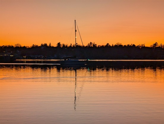 Original Bild:

Das Bild zeigt eine ruhige Wasserszene bei Sonnenuntergang. Im Vordergrund ist ein Boot mit einem Mast zu sehen, das auf dem ruhigen Wasser liegt. Die Wasseroberfläche spiegelt die warmen Farben des Himmels wider, der in einem sanften Orange- und Gelbtönen erstrahlt. Im Hintergrund ist eine Silhouette von Bäumen und Gebäuden zu erkennen, die den Horizont markieren. Die Szene ist von einer friedlichen Atmosphäre geprägt, die durch die sanften Wellen und die harmonische Farbgebung betont wird.