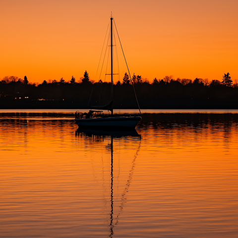 KI Bild:

Das Bild zeigt eine ruhige Szene am Abend, mit einem Segelboot im Vordergrund, das auf einer glatten Wasseroberfläche ruht. Der Himmel ist in warmen Orangetönen gefärbt, was auf einen Sonnenuntergang hinweist. Die Silhouette des Bootes, einschließlich seiner Masten und Seile, ist deutlich gegen den Hintergrund sichtbar. Im Hintergrund erstreckt sich eine Baumreihe, die den Horizont markiert. Die Wasseroberfläche spiegelt die Farben des Himmels und die Silhouette des Bootes wider, was eine friedliche und harmonische Atmosphäre schafft.