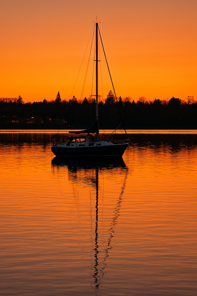 KI Bild:

Das Bild zeigt eine ruhige Szene am Abend, bei Sonnenuntergang. Im Vordergrund liegt ein Segelboot auf einer glatten Wasseroberfläche, dessen Silhouette sich deutlich im Wasser spiegelt. Der Mast des Bootes ragt hoch in den Himmel, und die Segel sind eingezogen. Der Himmel ist in warmen Orangetönen gefärbt, was auf den Sonnenuntergang hinweist. Im Hintergrund ist eine Baumgrenze zu sehen, die die Silhouette bildet, und es sind einige Lichter sichtbar, die auf Häuser oder Gebäude hinweisen. Die Wasseroberfläche spiegelt die Farben des Himmels wider, was eine friedliche und idyllische Atmosphäre schafft.