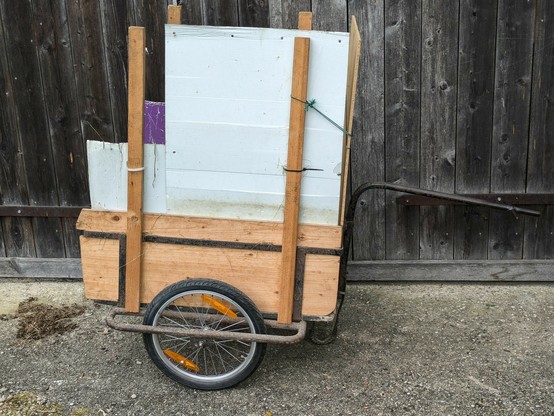 ein alter zweirädriger verrosteter Handkarren mit Deichsel und Holzplanken steht schief vor einem alten hölzernen Scheunentor. Das sichtbare Rad ist aus Alu und hat moderne orange Reflektoren drin