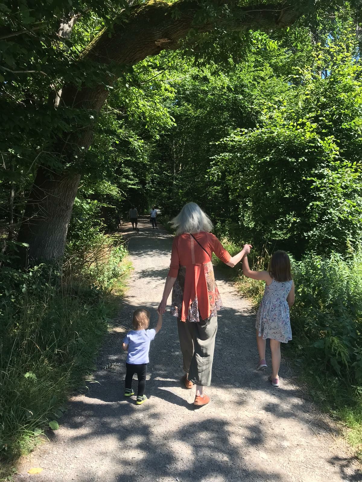 Auf dem Bild sieht man mich, die mit meinen zwei Enkelkindern einen Waldweg entlangspazieren. Ich halte die Kinder an beiden Händen – das kleinere Kind links trägt ein hellblaues T-Shirt und schwarze Hosen, das größere Kind rechts trägt ein sommerliches, geblümtes Kleid. Die Szene ist idyllisch: Wir befinden uns auf einem von Bäumen gesäumten Weg, durch den die Sonne hindurchscheint. Die Atmosphäre ist friedlich und familiär, ein typischer Sommertag im Grünen. Im Hintergrund sind noch weitere Spaziergänger auf dem Weg zu sehen.
Auf dem Bild sieht man mich, die mit meinen zwei Enkelkindern einen Waldweg entlangspazieren. Ich halte die Kinder an beiden Händen – das kleinere Kind links trägt ein hellblaues T-Shirt und schwarze Hosen, das größere Kind rechts trägt ein sommerliches, geblümtes Kleid. Die Szene ist idyllisch: Wir befinden uns auf einem von Bäumen gesäumten Weg, durch den die Sonne hindurchscheint. Die Atmosphäre ist friedlich und familiär, ein typischer Sommertag im Grünen. Im Hintergrund sind noch weitere Spaziergänger auf dem Weg zu sehen.