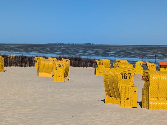 im Vordergrund gelbe Strandkörbe, dann schlickiges Watt und im Hintergrund zu erahnen eine kleine Insel 