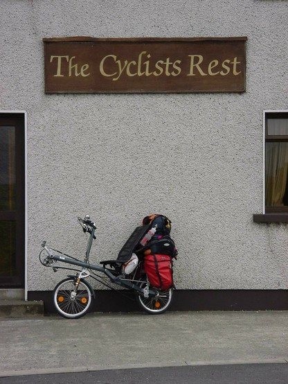 ein bepacktes kurzes Liegerad mit Obenlenker (Ostrad) parkt vor einem Gebäude, an dem ein Schild hängt The cyclists rest