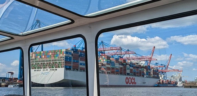 Blick durch die Fenster einer Barkasse auf 2 Container Schiffe. Das vordere grau (OOCL) und riesig und das hintere blaurostig und deutlich kleiner 