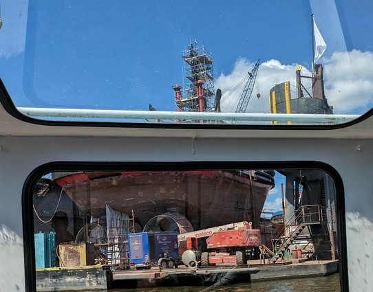 Blick durch die Fenster einer Barkasse auf das Heck eines Schleppers mit den beiden großen drehbaren Schrauben und Autoanhänger und fahrbarer Hebebühne 