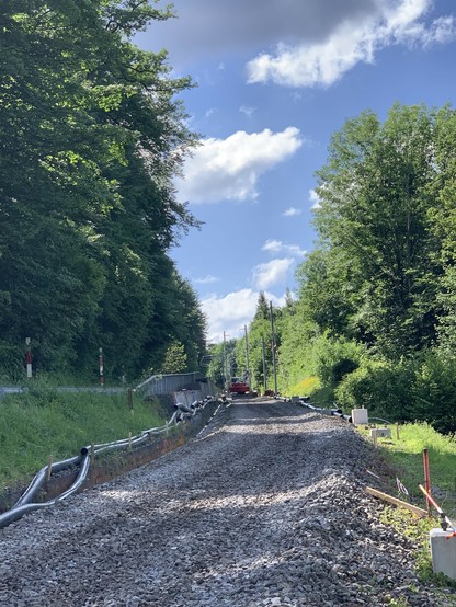 Blick bergwärts. Ein einsamer Bagger steht in einiger Entfernung auf den Schotterbett. Links und rechts hohe Bäume, der Himmel mit grossen Wolken, aber sonnig.