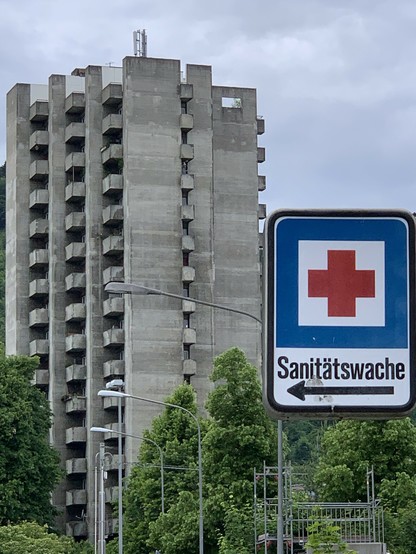 Ein hochkant aufgenommenes Bild mit dem besagten, herangezoomten Schild rechts unten im Vordergrund. Im unteren Hintergrund Baumbestand vor einem Hochhaus im Stil des Art Brut, das fast die ganze Höhe des Bildes einnimmt. Vom Vordergrund Richtung Hintergrund einige ebenfalls graue Peitschenkandelaber. Der graue Himmel passt dazu.